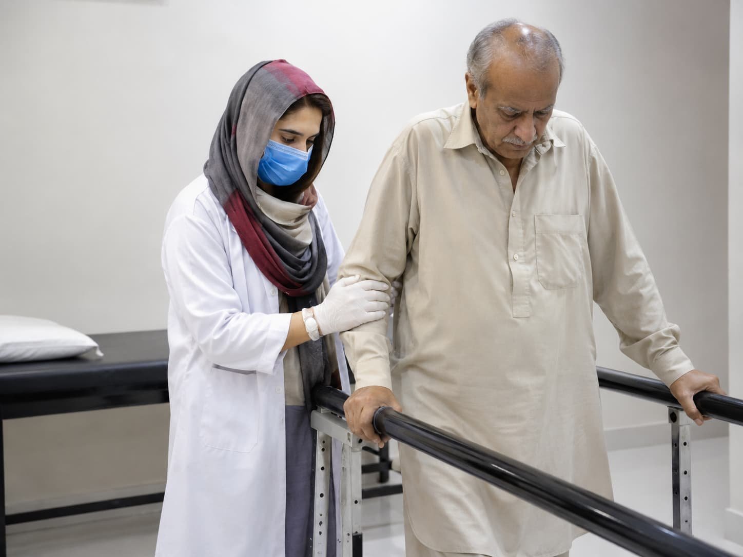 physiotherapist helping an older male patient walk using parallel bars during stroke rehabilitation